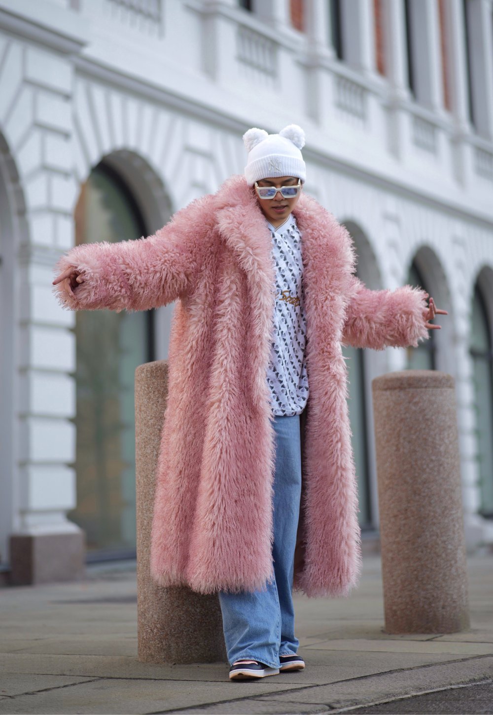 Casaco de pele falsa longo em rosa, com mangas largas, sobre uma camisa branca com padrões e jeans de ganga flare, acessorizado com um gorro branco.