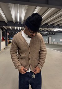 Άνδρας που φοράει μαύρο σκουφάκι, γυαλιά, μπεζ ζακέτα, λευκό πουκάμισο και σκούρο τζιν στέκεται κοιτάζοντας κάτω σε ένα αμυδρά φωτισμένο γκαράζ στάθμευσης.