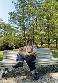 Πλεκτό πουλόβερ με σχέδιο φλόγας σε μπεζ και κόκκινο, συνδυασμένο με φαρδιά μπλε τζιν και μαύρα αθλητικά πα�πούτσια, καθισμένο σε έναν λευκό πάγκο.