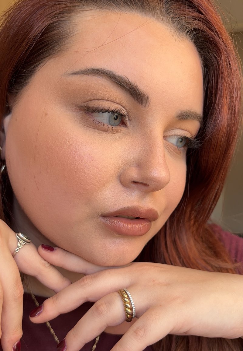 Nærbillede af et ansigt med mellemtonet hud, brunt hår, neutral makeup, sølv- og guldringe samt blødt konturerede ansigtstræk.