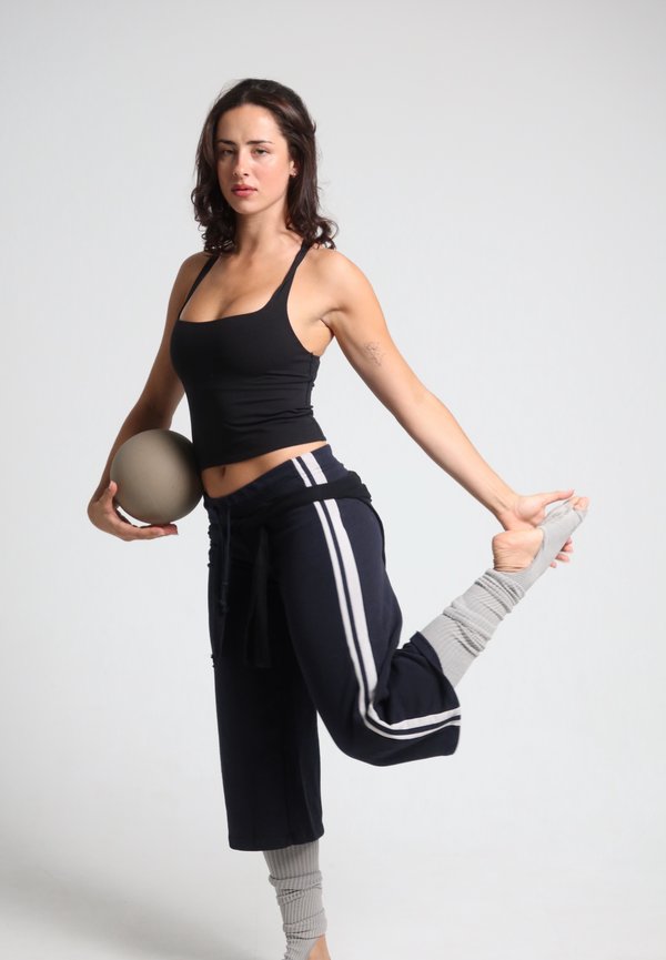 Mujer con camiseta sin mangas negra y pantalones azul marino estira la pierna hacia atrás mientras sostiene una pelota gris con una mano sobre un fondo liso.