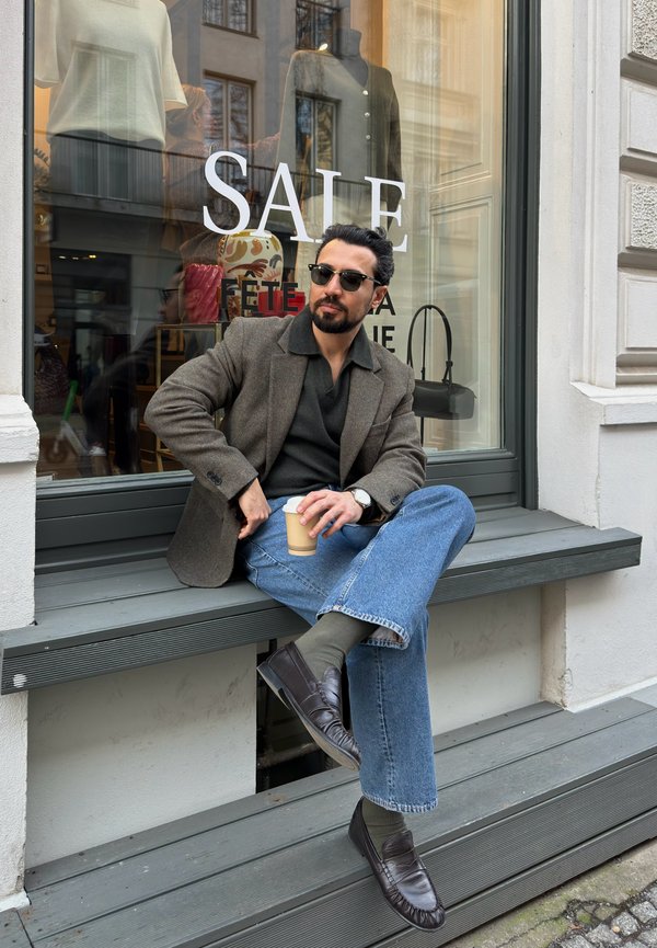 Homme portant des lunettes de soleil, un blazer marron et un jean, assis sur un banc en bois gris devant une vitrine de magasin avec un panneau "SOLDES", tenant une tasse de café.
