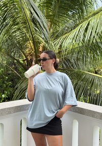 Hellblaues übergroßes T-Shirt, schwarze Minirock, hält einen cremefarbenen Trinkbehälter, umrahmt von üppigen grünen Palmblättern.