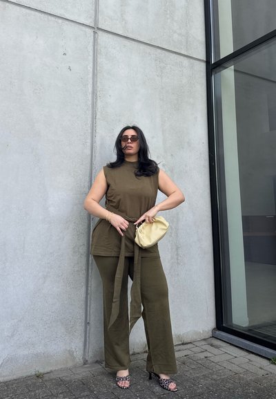Mujer con cabello largo y oscuro, vestida con top sin mangas verde oliva y pantalones, sosteniendo bolso beige, de pie junto a una pared de concreto cerca de una puerta de cristal.