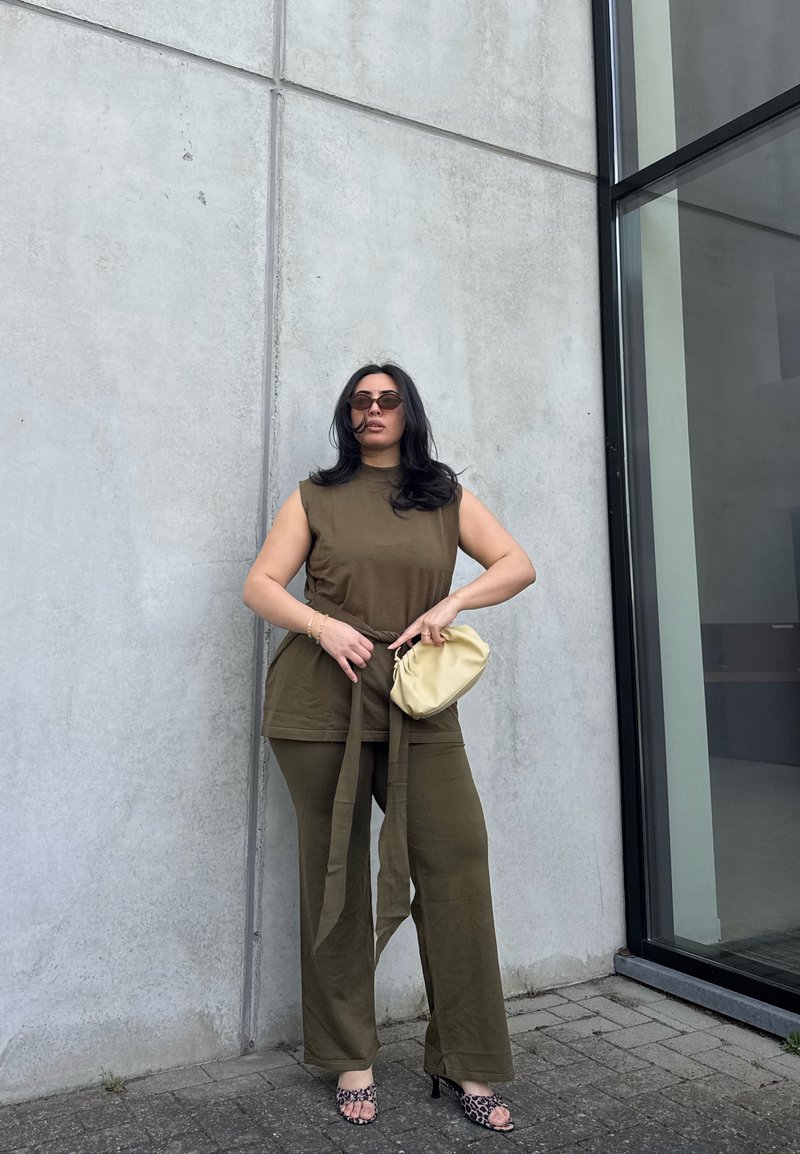 Woman with long dark hair wearing olive green sleeveless top and pants, holding beige purse, standing against concrete wall near glass door.