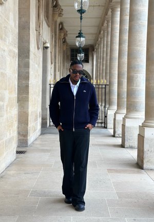 Hombre con gafas de sol y chaqueta azul marino caminando por un pasillo de columnas de piedra con faroles colgantes y una puerta de hierro al fondo.