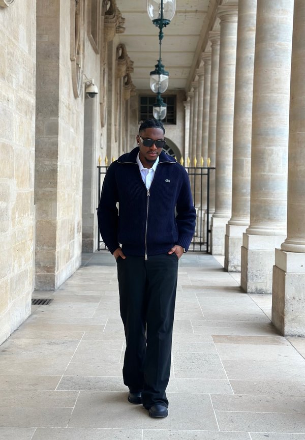 Homme portant des lunettes de soleil et une veste bleu marine marchant dans un couloir de colonnes en pierre avec des lanternes suspendues et une grille en fer en arrière-plan.