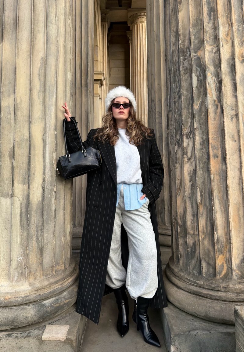 Une femme pose devant des colonnes en pierre, portant un manteau noir à rayures, un sweat-shirt gris, une chemise bleu clair, un pantalon gris et des bottes noires à bout pointu.