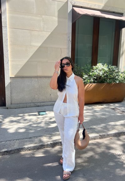 Conjunto de dos piezas de lino blanco que incluye una blusa sin mangas con cuello y un corte frontal, junto con pantalones holgados. Bolso marrón y sandalias.