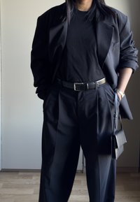 Blazer noir sur un t-shirt noir, assorti à un pantalon noir ample. Comprend une ceinture noire brillante et un petit sac à main noir. Textures lisses.