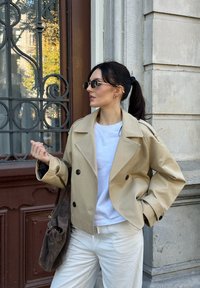 Béžové dvojradové sako s veľkými gombíkmi, nosené na bielu košeľu. Doplnky zahŕňajú okrúhle slnečné okuliare a tmavohned�ú tašku.