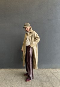 Beige blankregnfrakk, kremgenser, brune joggebukser med rosa striper, og bordeaux sneakers. En grønn caps gir kontrast til antrekket.