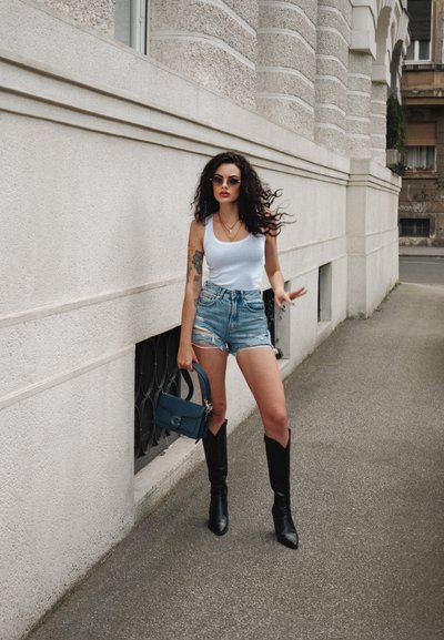 La modelo lleva una blusa blanca sin mangas, pantalones cortos de denim desgastados y botas negras hasta la rodilla, sosteniendo un bolso azul contra una pared texturizada.