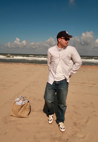 Camisa de color claro, abotonada, sobre una camiseta blanca, jeans azules y zapatillas marrones. Una bolsa marrón con ropa doblada está sobre la arena.