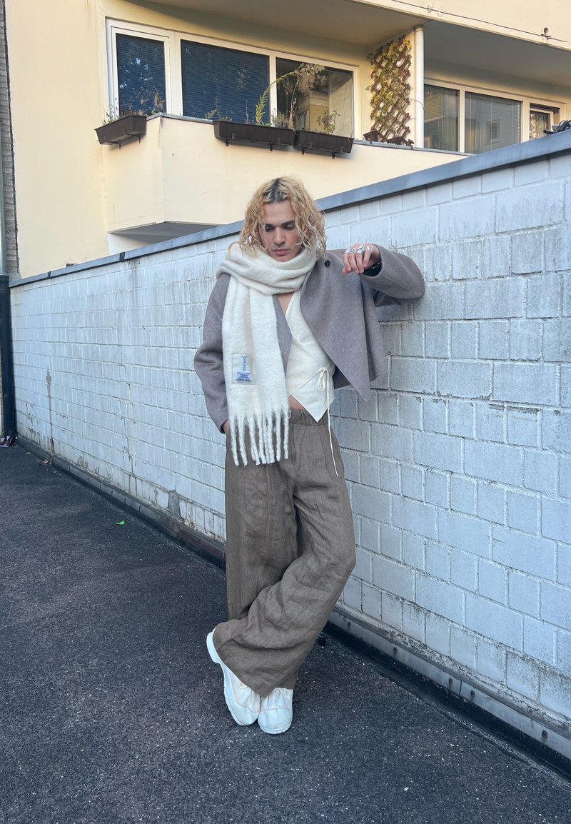 Beige wollen jas met een cropped ontwerp, witte sjaal met franjes, crème top, bruine linnen broek en witte schoenen. Stedelijke setting met een grijze muur.