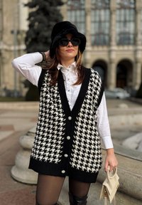 Czarnobiały sweter w wzór houndstooth z głębokim dekoltem w serek, ozdobnymi guzikami, zestawiony z białą koszulą i kapeluszem.