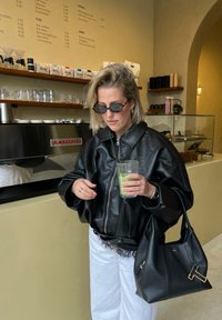 Veste oversize en cuir noir, pantalon blanc à jambes larges, sac à main noir avec des accents dorés, et une boisson verte dans un verre.