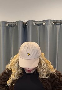 Casquette de baseball beige en tissu, ornée d'un petit logo papillon noir et jaune à l'avant. Portée avec une chemise à carreaux marron.