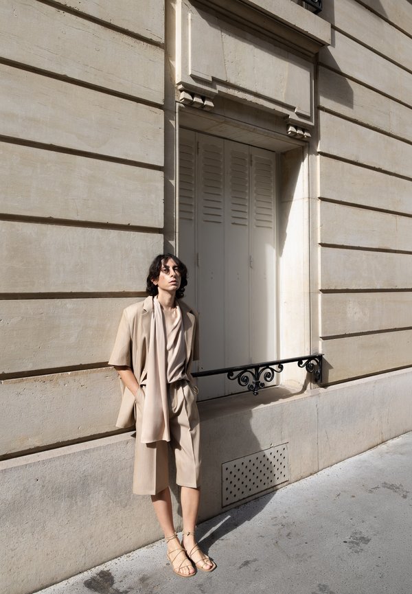 Persona con ropa beige y sandalias apoyada contra una pared de piedra beige de un edificio con contraventanas cerradas y una barandilla metálica decorativa.