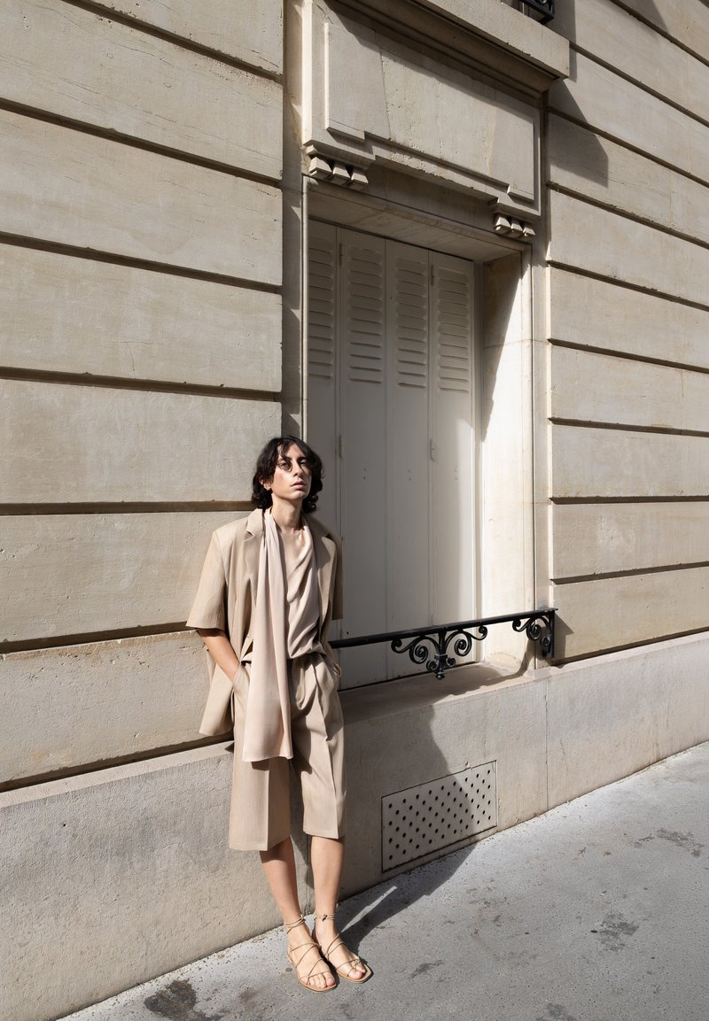 Persona con ropa beige y sandalias apoyada contra una pared de piedra beige de un edificio con contraventanas cerradas y una barandilla metálica decorativa.