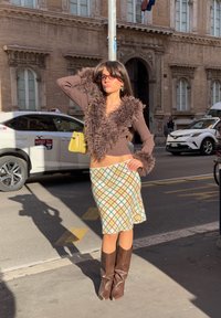 Braun besetzter, kurz geschnittener Pullover mit Fellbesatz, gemusterter Rock in Grün, Gelb und Braun, braune kniehohe Stiefel, gelbe Handtasche, städtische Straßenszene.