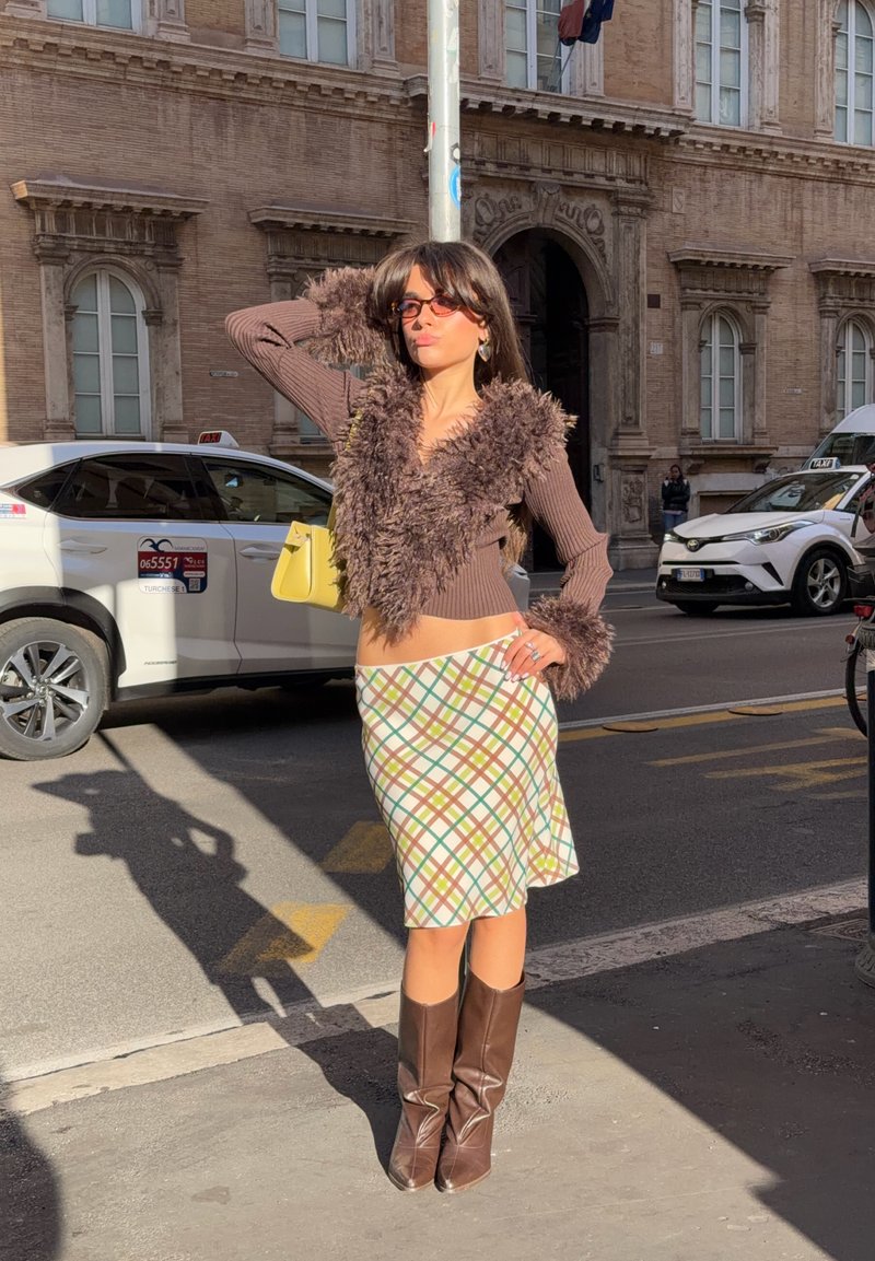Maglione corto con bordo in pelliccia marrone, gonna con motivo verde, giallo e marrone, stivali marroni al ginocchio, borsa gialla, scena urbana di strada.