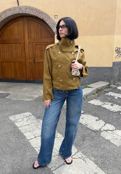 Mujer con corte de pelo bob negro que lleva una chaqueta marrón de doble botonadura, vaqueros anchos azules, chanclas, gafas de sol y sostiene un pequeño bolso tejido.