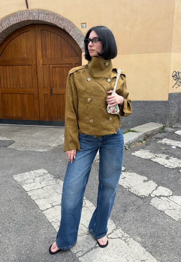 Femme avec une coupe au carré noire portant une veste marron croisée, un jean large bleu, des tongs, des lunettes de soleil et tenant un petit sac tissé.