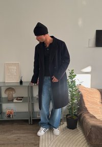 Homme en manteau noir, bonnet et baskets, debout près d'une étagère en métal avec des décorations et une plante en pot à côté d'un canapé en velours côtelé marron dans une pièce lumineuse.