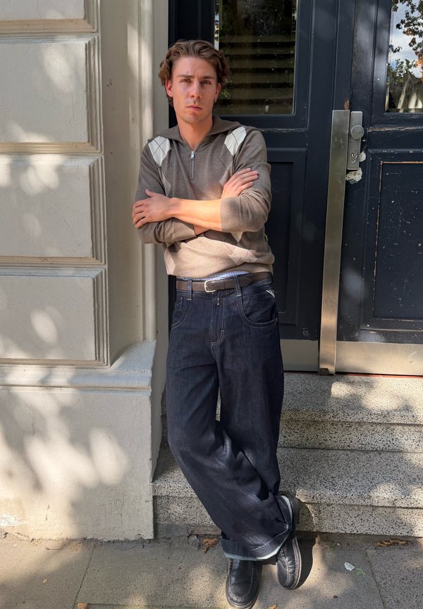 Jeune homme aux bras croisés appuyé contre un mur près d'une porte sombre, portant un pull marron et un jean foncé, sous la lumière tachetée du soleil.