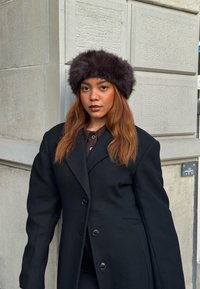 Svart oversized kappa med stora ärmar, brun pälsmössa och en mörk topp, mot en strukturerad stenvägg som bakgrund.