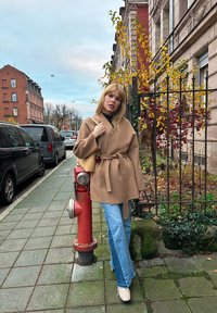 Manteau beige avec de longues manches, ceinturé à la taille, associé à un jean flair bleu clair et des chaussures beiges. Fond urbain avec un poteau de réservoir d'incendie.