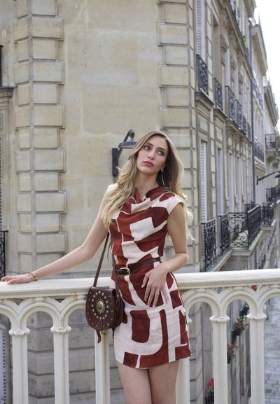 Mujer con un vestido rojo y blanco con estampado se apoya en una barandilla de piedra blanca en una calle parisina con edificios históricos al fondo.