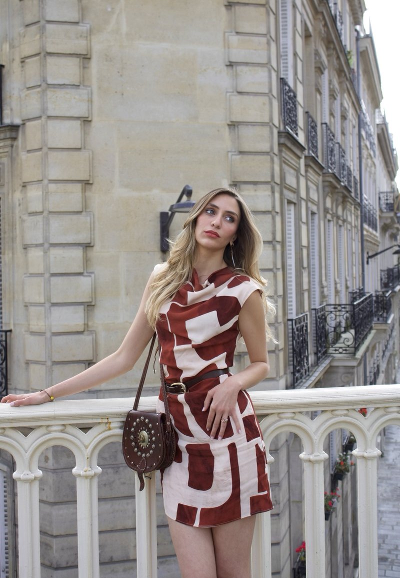 Femme vêtue d'une robe rouge et blanche à motifs, appuyée sur une balustrade en pierre blanche dans une rue parisienne avec des bâtiments historiques en arrière-plan.