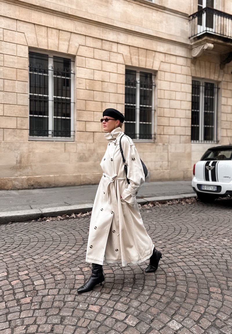 Femme en long trench-coat beige, béret noir et bottes marchant sur une rue pavée avec un bâtiment en pierre et une voiture garée en arrière-plan.