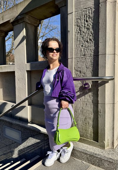 Rompevientos morado, camiseta blanca, falda de canalé lavanda, zapatillas blancas y bolso verde. Texturas suaves con un contraste de colores brillantes.