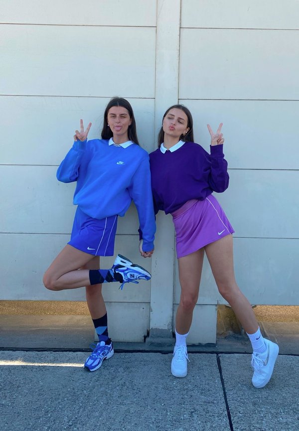 Dos personas están juntas, una vistiendo un sudadera azul claro y una falda morada, y la otra con un sudadera morada oscuro y una falda rosa, ambas con zapatillas.