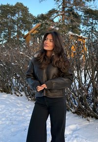 Femme en veste et pantalon en cuir noir debout dans la neige près de buissons dépouillés, avec une maison jaune et des pins en arrière-plan.