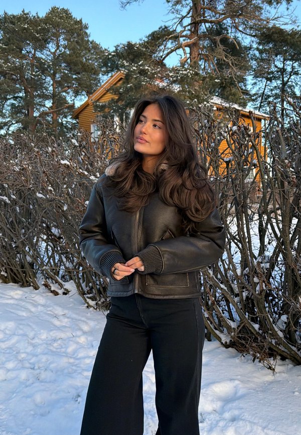 Kvinne i svart skinnjakke og bukser står i snøen nær nakne busker med et gult hus og furutrær i bakgrunnen.