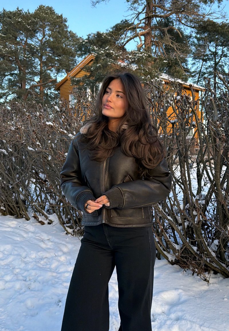 Femme en veste et pantalon en cuir noir debout dans la neige près de buissons dépouillés, avec une maison jaune et des pins en arrière-plan.