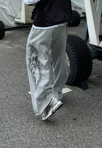 Γκρι φόρμες με γραφικό αστέρι στο αριστερό πόδι, συνδυασμένες με ασημένια αθλητικά παπούτσια. Στο βάθος περιλαμβάνεται ένα καρότσι με ρόδες και ασφάλτινη επιφάνεια.