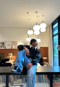 En person med svart topp og rivne jeans sitter på en bokhylle i et moderne rom med hengende globelys og innrammet kunst.