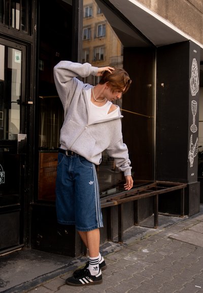 Sudadera gris con cremallera, camiseta sin mangas blanca, pantalones cortos de mezclilla azules con un diseño de tres rayas de Adidas, zapatillas negras, de pie cerca de una tienda.