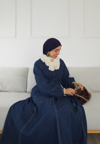 Vestido largo de mezclilla con un cuello de crema con volantes, combinado con una bufanda negra y accesorios dorados, sentado en un sofá de tonalidades claras.