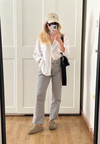 Camisa blanca de gran tamaño con botones, jeans de talle alto grises, gorra beige y zapatos verdes tipo slip-on. Bolso negro bandolera con correa. Fondo de suelo de madera.