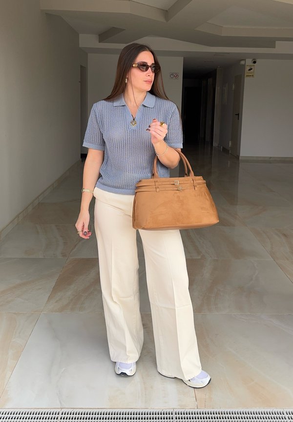 Woman wearing sunglasses, a blue knitted polo shirt, white trousers, and sneakers, holding a large brown handbag, standing in a tiled indoor corridor.