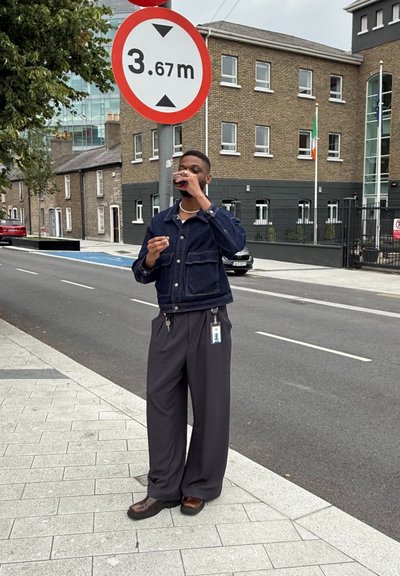 Chaqueta de mezclilla oscura sobre una camisa blanca, pantalones negros holgados y zapatos marrones. Sosteniendo una bebida, de pie junto a un cartel de restricción de altura.