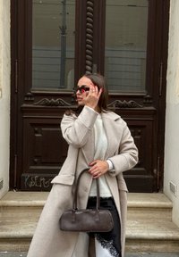 Femme en manteau beige et pull blanc ajustant ses lunettes de soleil, tenant un petit sac à main foncé, debout devant une porte en bois sculptée.