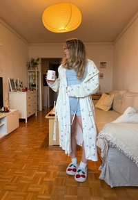 Peignoir blanc à motifs d'oiseaux, porté sur un haut bleu clair et un short. Chaussons blancs avec motif cerise. Intérieur chaleureux avec un sol en bois.