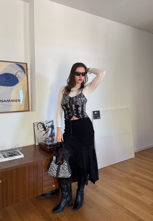 Femme portant des lunettes de soleil noires, un top en dentelle sur un t-shirt blanc à manches longues, une jupe noire et des bottes, tenant un sac à motif zèbre dans une pièce moderne avec des œuvres d'art.